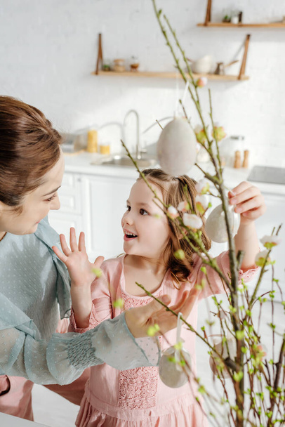 selektivní zaměření překvapeného dítěte při pohledu na matku v blízkosti dekorativních velikonočních vajec na vrbových větvích  - Fotografie, Obrázek