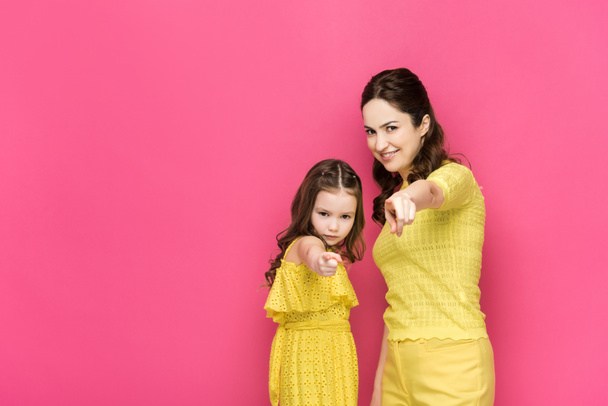 serious kid and happy mother pointing with fingers isolated on pink  - Фото, изображение