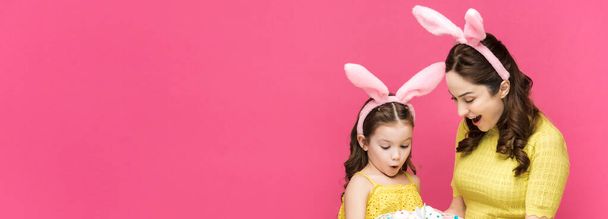 plano panorámico de madre e hija sorprendidas en orejas de conejo mirando huevos de Pascua aislados en rosa
  - Foto, imagen