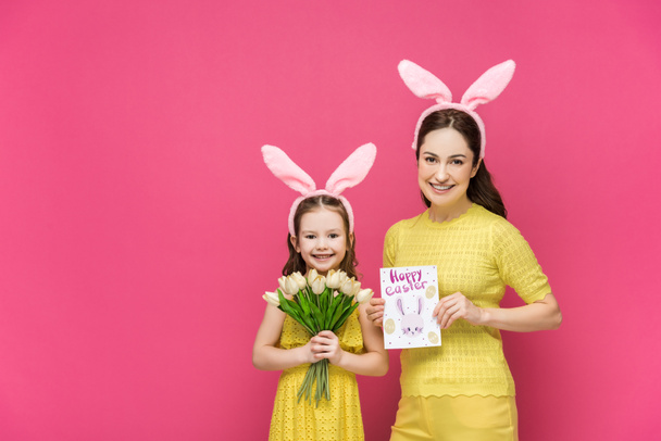 ευτυχισμένη μητέρα στα αυτιά λαγουδάκι κρατώντας ευχετήρια κάρτα με χαρούμενα γράμματα Πάσχα κοντά κόρη με τουλίπες απομονωμένες σε ροζ  - Φωτογραφία, εικόνα