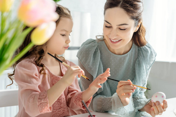 selective focus of happy mother and daughter painting chicken egg near tulips  - Photo, Image