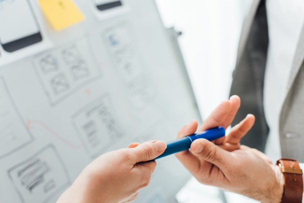 Cropped view of ux designer giving marker to colleague near project of ux design on whiteboard on white background - 写真・画像