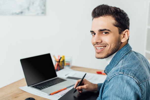 Usmívající se ux návrhář při pohledu na fotoaparát při použití grafického tabletu a notebooku u stolu  - Fotografie, Obrázek