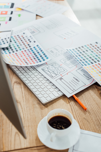 Selektivní zaměření šálku kávy a monitoru v blízkosti barevných palet a náčrtků designu uxu na dřevěném stole - Fotografie, Obrázek