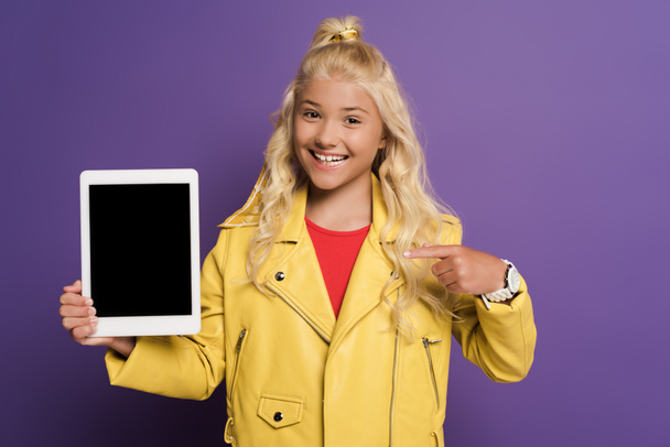Lächelndes Kind zeigt mit dem Finger auf digitales Tablet auf lila Hintergrund  - Foto, Bild