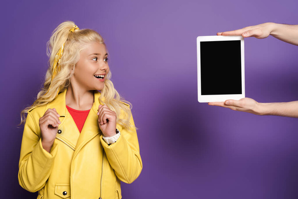 紫の背景に驚く子供にデジタルタブレットを与える男のクロップドビュー  - 写真・画像