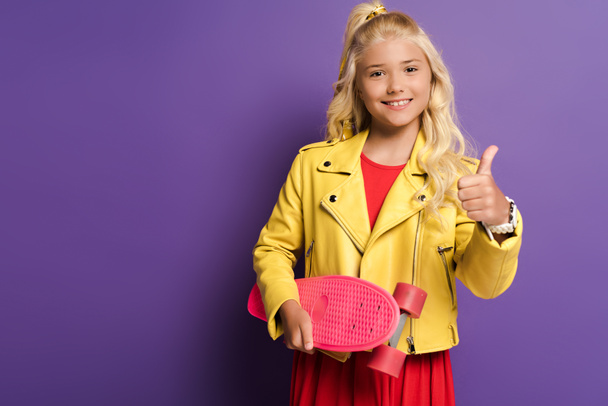 sonriente niño sosteniendo penny board y mostrando como en púrpura fondo
  - Foto, Imagen