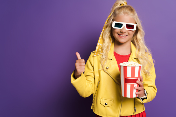 smiling kid with 3d glasses holding bucket with popcorn and showing like on purple background  - Photo, Image