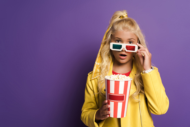 shocked kid with 3d glasses holding bucket with popcorn on purple background  - Photo, Image