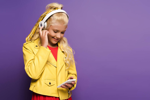 glimlachend kind met hoofdtelefoon met behulp van smartphone op paarse achtergrond  - Foto, afbeelding