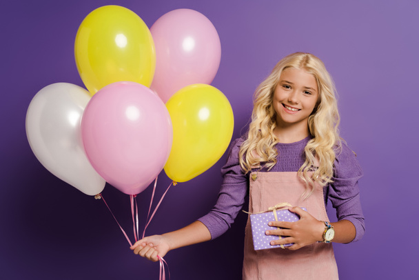 smiling kid holding balloons and gift box on purple background  - Foto, Imagem