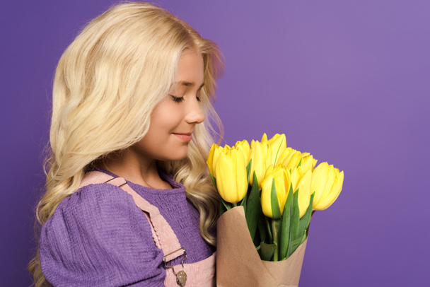 side view of smiling kid holding bouquet of tulips on purple background  - Foto, imagen