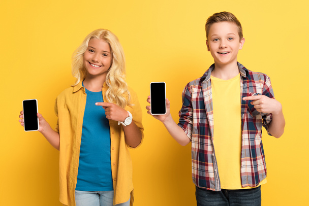 smiling kids pointing with fingers at smartphones on yellow background  - Zdjęcie, obraz