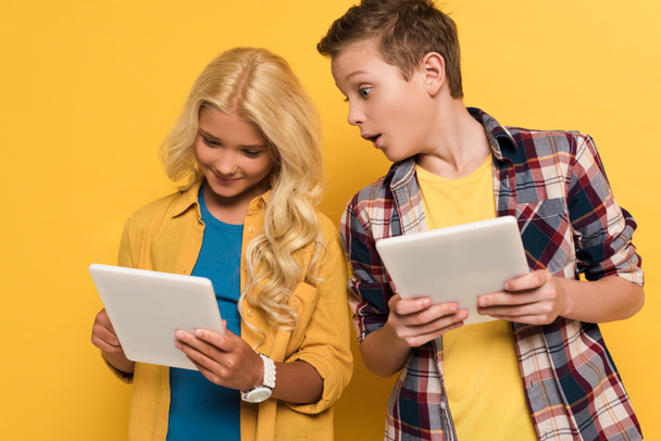 shocked kid looking at digital tablet of his smiling friend on yellow background  - Fotó, kép