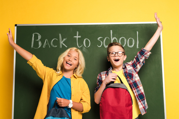 low angle view of smiling schoolkids with outstretched hands holding backpacks and standing near chalkboard with back to school lettering  - Foto, imagen