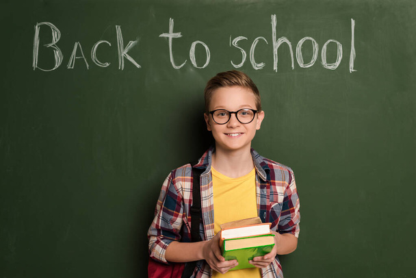 smiling schoolboy with books standing near chalkboard with back to school lettering - Φωτογραφία, εικόνα