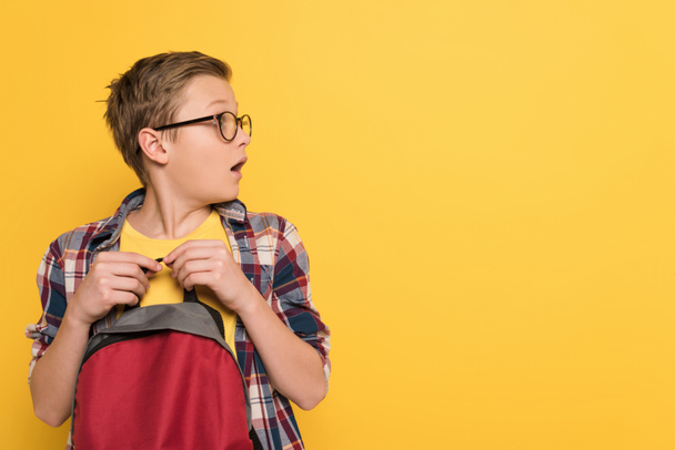 shocked schoolboy with glasses holding backpack isolated on yellow - Photo, image