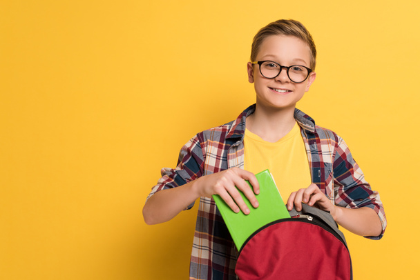 sonriente escolar poniendo libro en mochila sobre fondo amarillo
  - Foto, Imagen