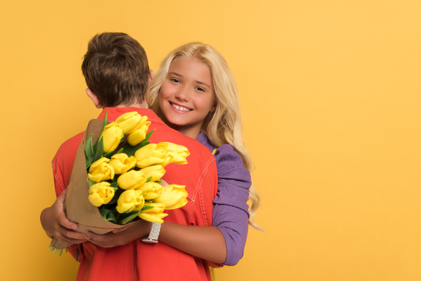 back view of boy hugging smiling friend with bouquet on yellow background  - Zdjęcie, obraz