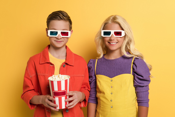 3Dメガネをかけた笑顔の子供たちが黄色の背景にポップコーンを持ち  - 写真・画像