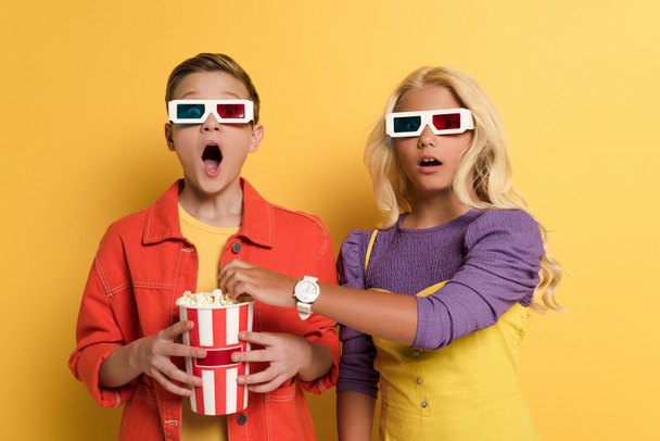shocked kids with 3d glasses holding popcorn and looking at camera on yellow background  - Photo, Image