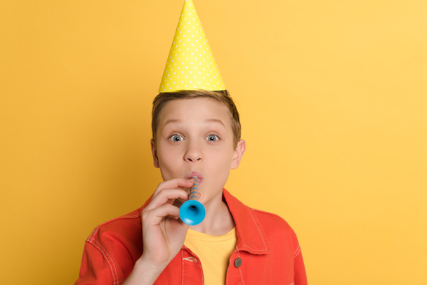 Kind in Partymütze bläst in Partyhorn auf gelbem Hintergrund  - Foto, Bild