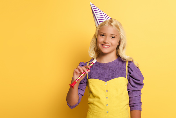 glimlachend kind met party cap holding party hoorn op gele achtergrond  - Foto, afbeelding