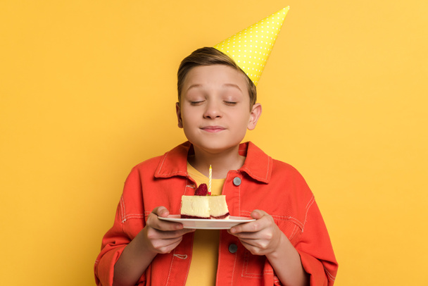 smiling kid with closed eyes making wish and holding plate with birthday cake on yellow background  - Fotoğraf, Görsel
