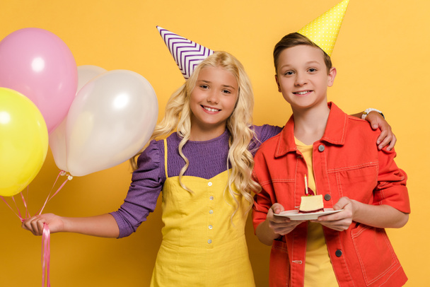 smiling kids with party caps holding balloons and plate with birthday cake on yellow background  - Foto, afbeelding