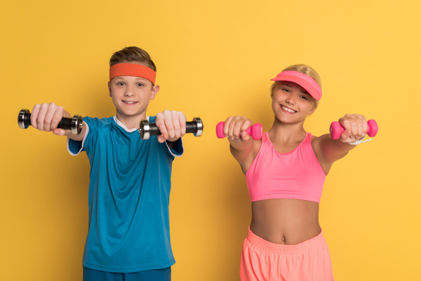 smiling kids in sportswear training with dumbbells on yellow background  - Foto, Imagem