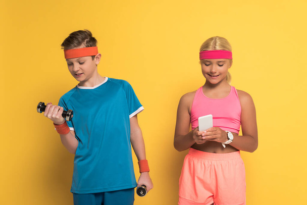 smiling boy training with dumbbells and his friend  holding smartphone on yellow background  - Photo, Image