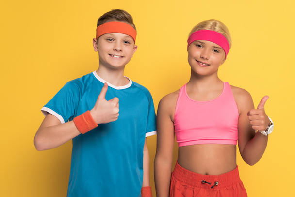 Lächelnde Kinder in Sportbekleidung zeigen Daumen hoch auf gelbem Hintergrund  - Foto, Bild