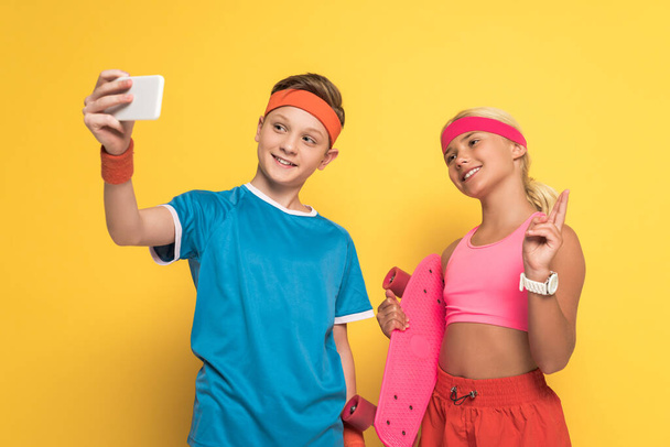 smiling boy taking selfie and his friend holding penny board and showing peace gesture on yellow background  - Photo, Image