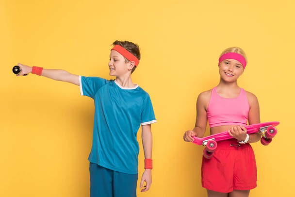 sorrindo menino treinamento com halteres e seu amigo segurando penny board no fundo amarelo
  - Foto, Imagem