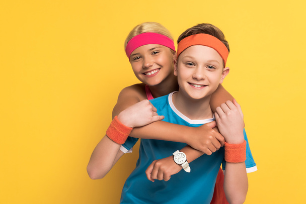 Lächelnde Kinder in Sportkleidung umarmen sich und schauen in die Kamera auf gelbem Hintergrund  - Foto, Bild