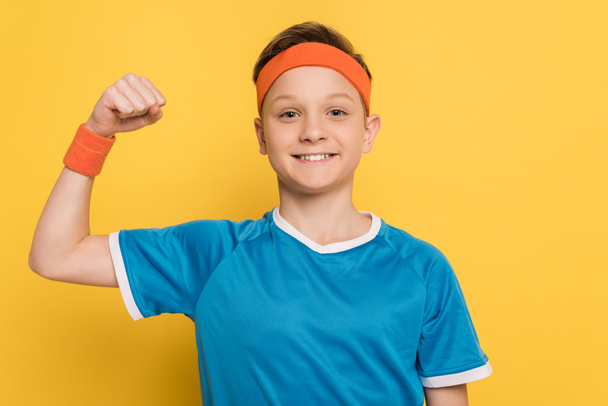 χαμογελαστό παιδί σε αθλητικά ρούχα δείχνει ισχυρή χειρονομία σε κίτρινο φόντο  - Φωτογραφία, εικόνα