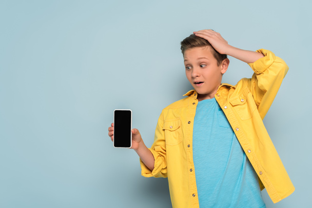 shocked kid touching head and holding smartphone on blue background  - Fotó, kép