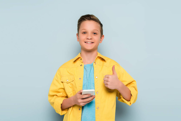 smiling kid holding smartphone and showing like on blue background  - Foto, imagen
