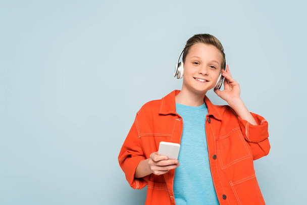 smiling kid with headphones listening to music and holding smartphone on blue background  - Fotó, kép