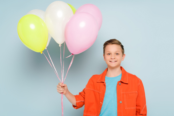 smiling kid holding balloons and looking at camera on blue background  - Φωτογραφία, εικόνα