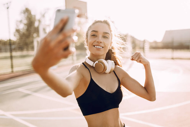 Beautiful fitness woman wearing headphones taking selfie and showing her muscles outdoors - Photo, image