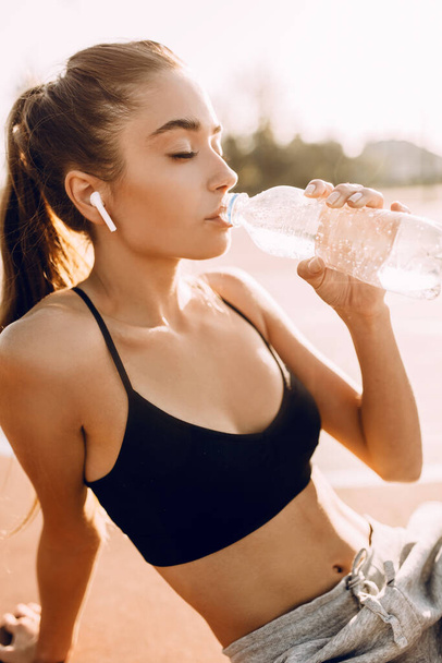 Sporty young girl, in top and sweatpants, drinking water from a bottle, sitting on the field after a workout - Foto, Imagen