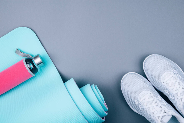 Top view of white sport shoes sneakers, blue green yoga mat and reusable glass water bottle on pastel grey background. Yoga pilates or fitness practice. Losing weight and sport concept. Flat Lay. - Photo, Image