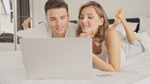 An attractive smiling young couple man and woman are using the laptop computer in a white bedroom in the morning - Filmati, video