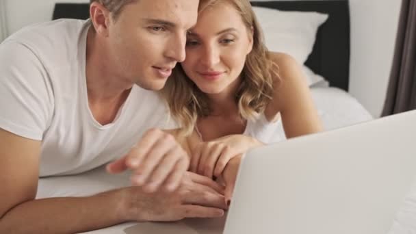 A lovely young couple are watching something on the laptop computer while lying in bed in a white bedroom in the morning - Filmati, video