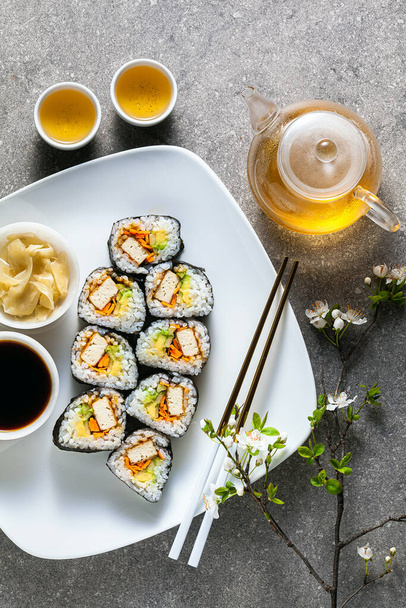 vegan sushi with tofu in General Tso sauce, avocado and fresh vegetables . on a table with branches of a blossoming tree and green tea in a glass teapot - Fotografie, Obrázek