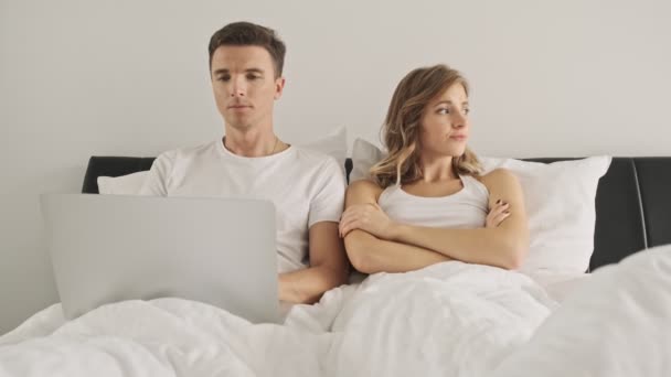 A handsome boy is using a laptop computer while his girlfriend is sitting with crossed hands in a white bedroom in the morning - Filmati, video