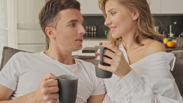 Attractive young couple man and woman in casual wear drinking tea at home while sitting on sofa in living room - Záběry, video