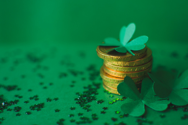 Saint Patricks day backdrop with close up stack of chocolate coins with green four-leafed paper shamrocks on green blurred background with small stars. Lucky concept. Copy space. Selective focus - Zdjęcie, obraz