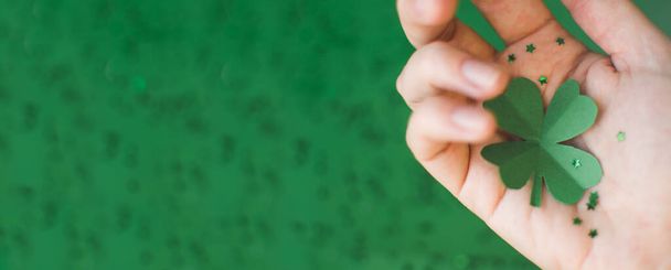 Πανό ημέρας του Αγίου Πατρικίου με κορυφαία θέα του πράσινου τετράφυλλου τριφύλλι χαρτί σε γυναίκες χέρι πασπαλισμένο με μικρά αστέρια σε πράσινο θολή φόντο. Τυχερή ιδέα. Χώρος για κείμενο. Επιλεκτική εστίαση - Φωτογραφία, εικόνα